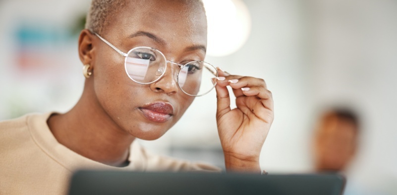 Portrait of a woman adjusting her glasses while working on a laptop, focusing on the screen in a bright, modern workspace.