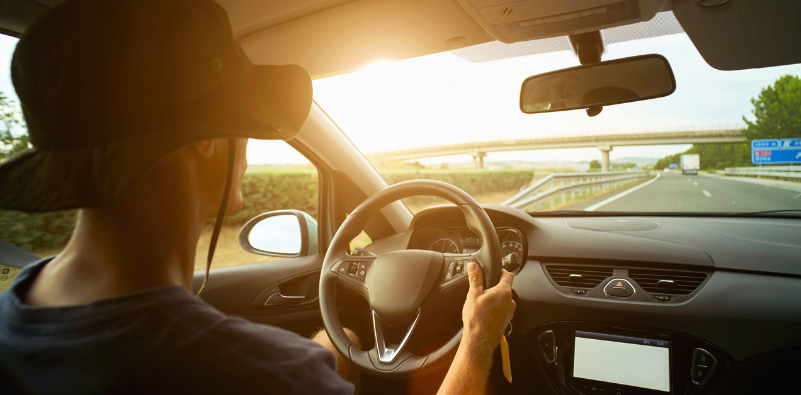 Driver wearing a hat on a sunny day, seen from behind inside a car