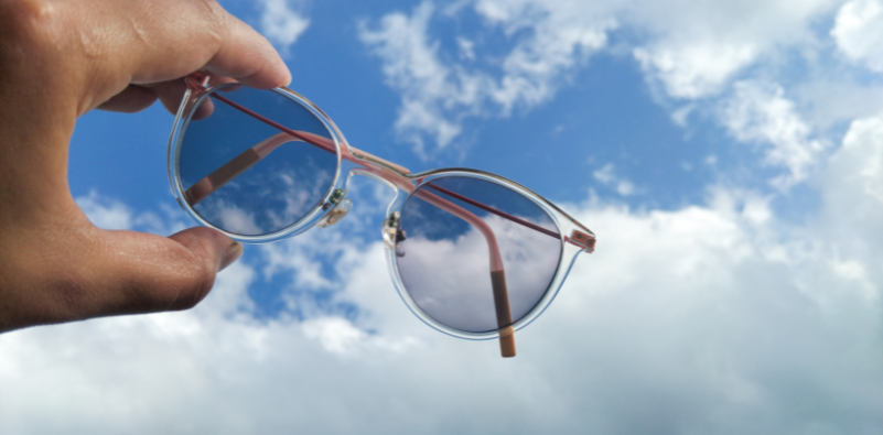 Hand holding semi-darkened photochromic glasses up to the blue sky with clouds.